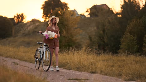 Joven-Hermosa-Rubia-En-Verano-Con-Vestido-Y-Sombrero-Caminando-Por-La-Carretera-Con-Bicicleta-Y-Flores-En-Cámara-Lenta.